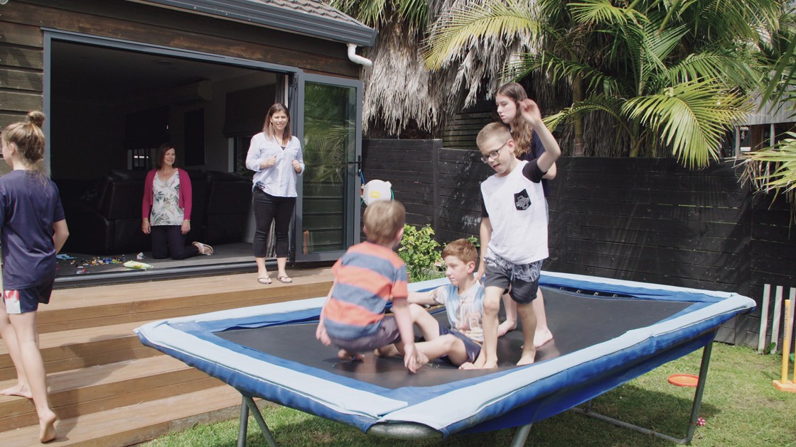 Four children play on a trampoline supervised by parents at a house. 