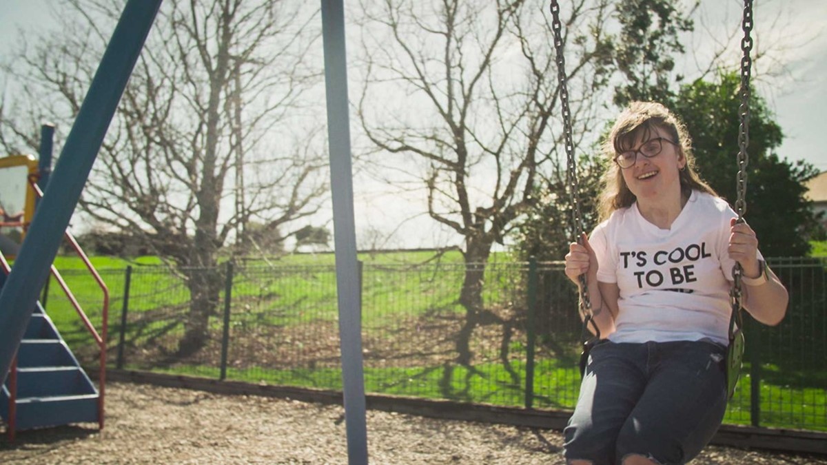 Sarah sitting on a swing in a playground, wearing a shirt that says; "It's cool to be kind"
