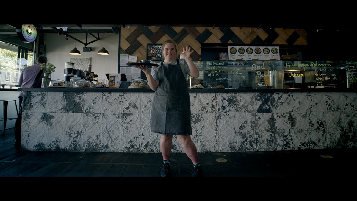 Emma stands in a cafe holding her serving tray and waving